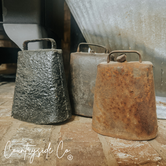 Three rustic metal cowbells of different sizes on a stone surface with 'Countryside Co' branding.